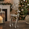 Silver nickel-plated aluminium standing stag figurine with branching antlers styled on a dark wood table beside a lit lantern with Christmas tree in background
