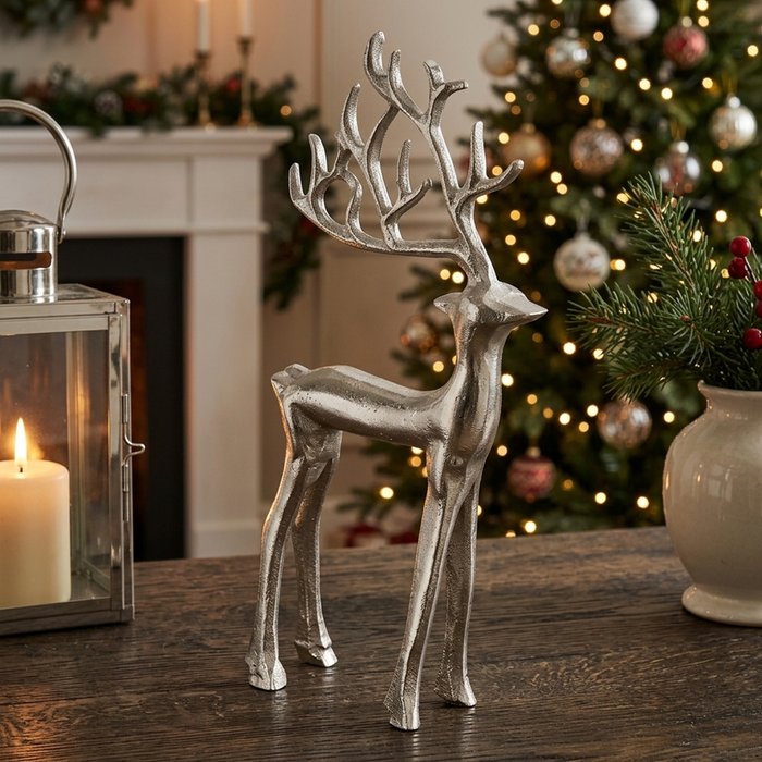 Silver nickel-plated aluminium standing stag figurine with branching antlers styled on a dark wood table beside a lit lantern with Christmas tree in background