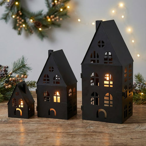 Set of three matte black metal house lanterns with tealights glowing through cut-out windows, styled with Christmas foliage