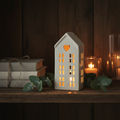 Dainty white ceramic LED house glowing warmly on a dark wooden shelf in a cosy Nordic hygge setting